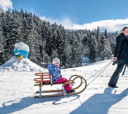 Mutter mit Baby im Tragetuch zieht ihre Tochter am Schlitten nach