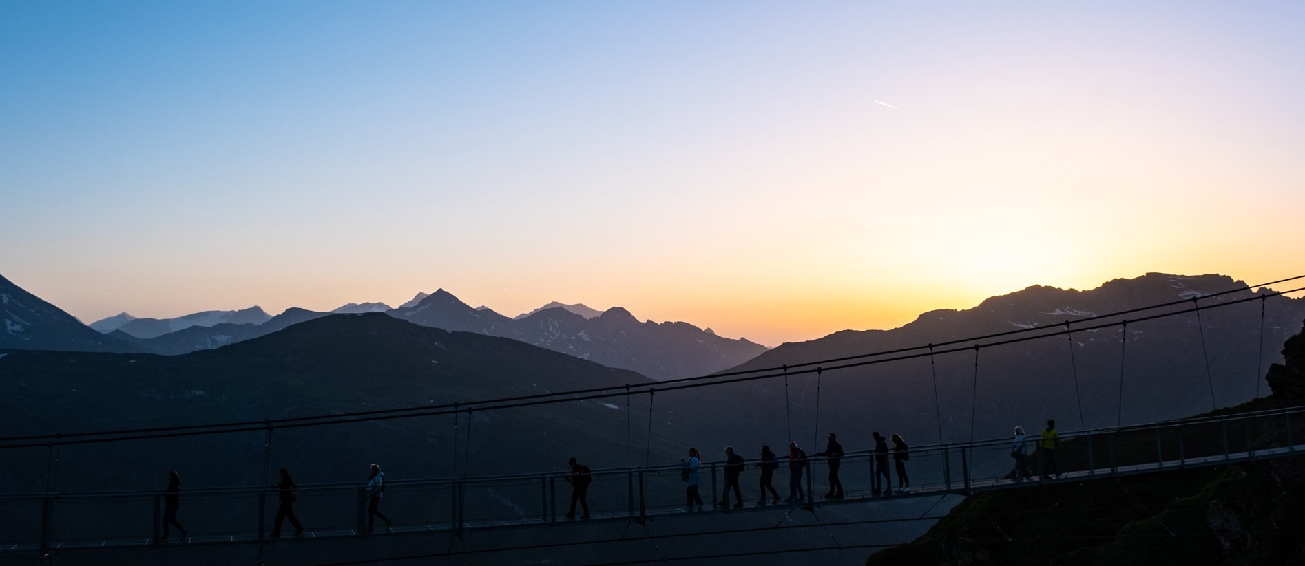 Dieses Bild zeigt die Hängebrücke auf dem Stubnerkogel bei Sonnenuntergang. Eine Gruppe läuft hinüber.
