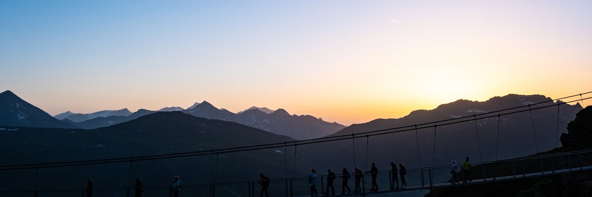 Dieses Bild zeigt die Hängebrücke auf dem Stubnerkogel bei Sonnenuntergang. Eine Gruppe läuft hinüber.