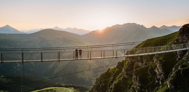 Dieses Bild zeigt die Hängebrücke am Stubnerkogel im Sommer mit zwei Menschen bei Sonnenuntergang