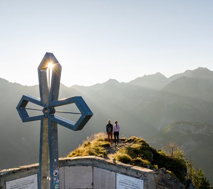 Dieses Bild zeigt zwei Frauen, die vor dem Gipfelkreuz des Hüttenkogels stehen und den Sonnenuntergand auf dem Berg genießen.